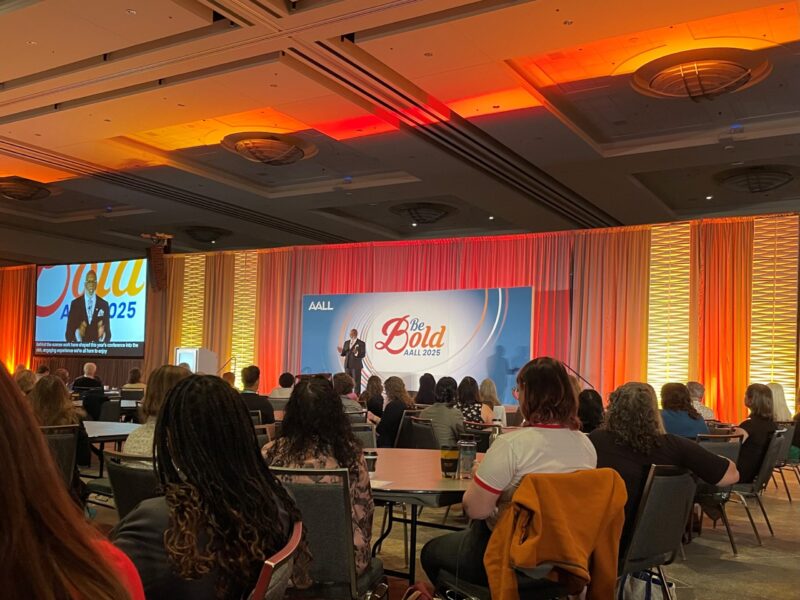 The opening keynote at the AALL annual meeting. A ballroom lit up in bright orange, red and yellow lights. Attendees are seated at round tables. the AALL President, Cornell Winston, is on stage in front of a projector with the conference theme, Be Bold in large letters.