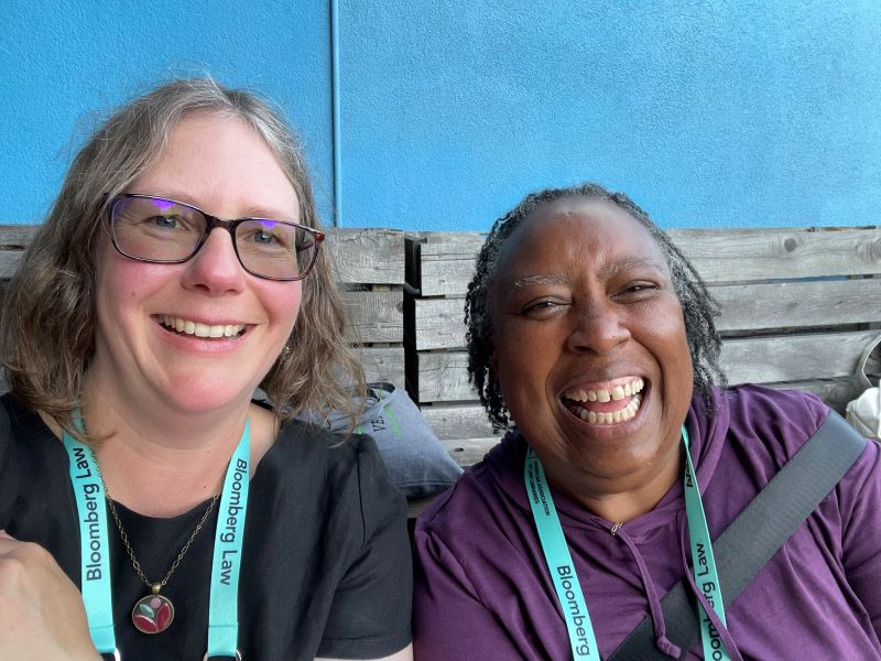 Sarah, a white lady with glasses, on the left, and Eli, a Black lady wearing a purple shirt on the right. Both have big smiles on their faces.
